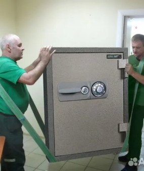 Двое профессиональных грузчиков в зеленой униформе перемещают массивный сейф с помощью специальных ремней, демонстрируя услугу безопасного и аккуратного перемещения крупногабаритных и тяжелых предметов.