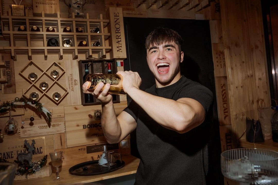 Бармен с улыбкой энергично встряхивает шейкер в уютном деревянном баре с винными полками на заднем плане