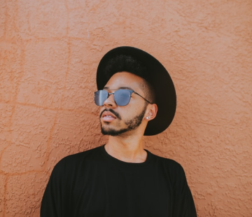 Парень в очках, чёрной шляпе и черной футболке на фоне грязно-розовой стены.